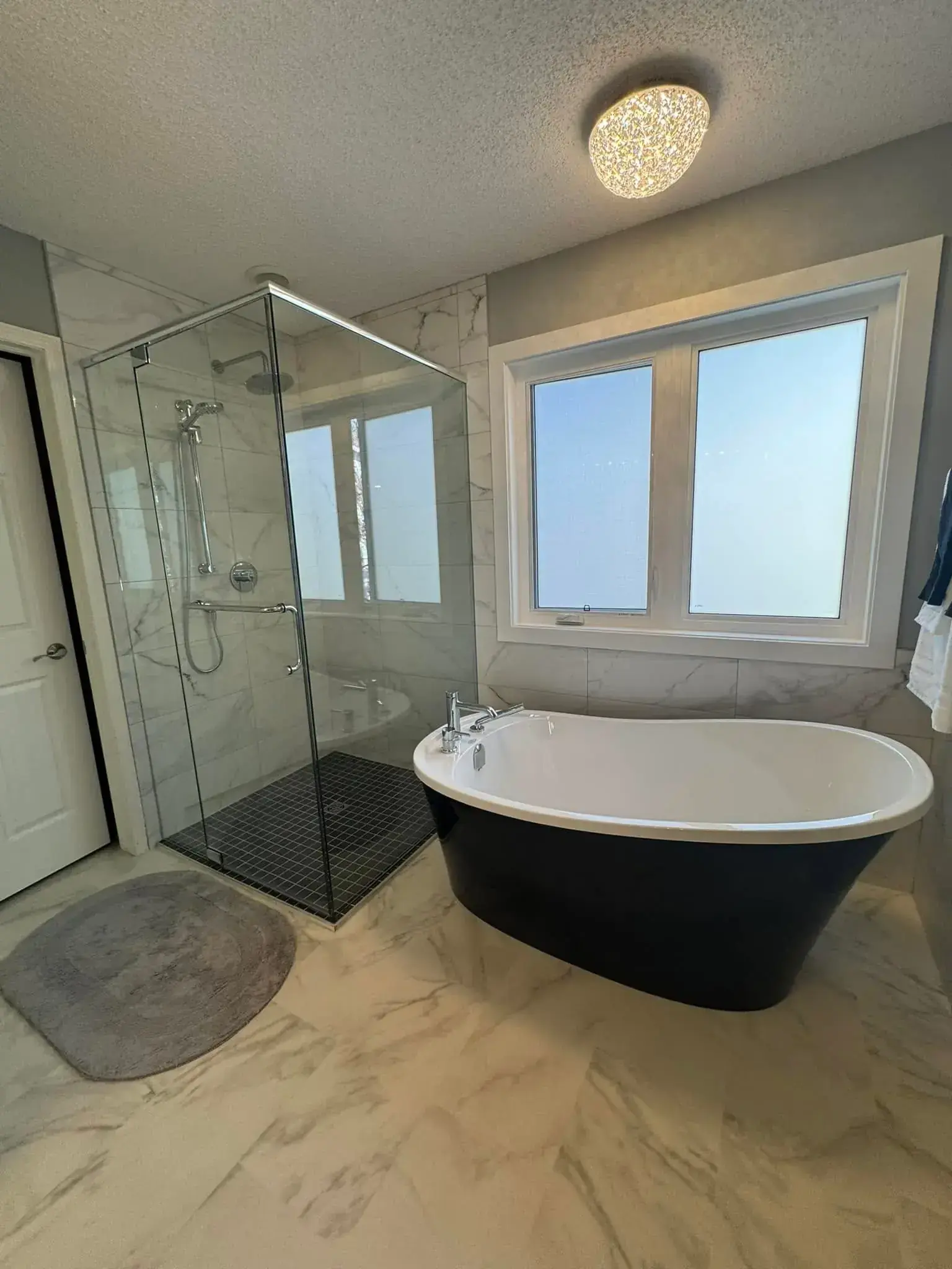 Black freestanding soaker tub with frameless glass shower enclosure, marble-look tile, and crystal ceiling light in Calgary
