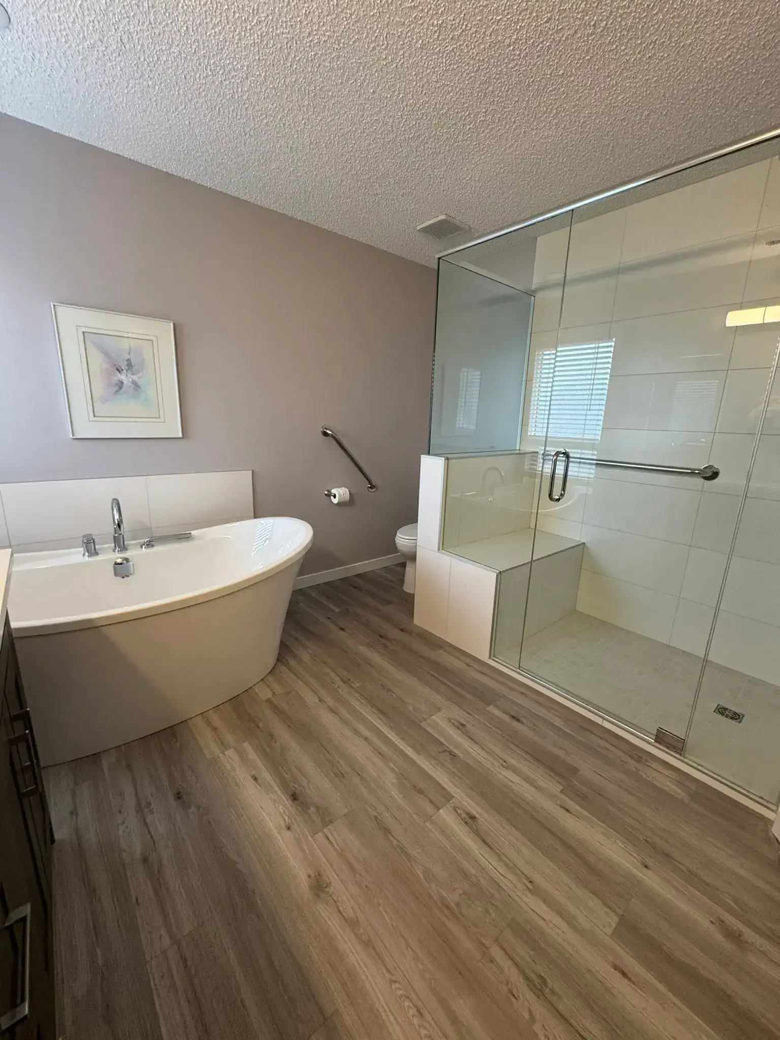 Freestanding white tub with large glass walk-in shower, built-in bench, grab bar, and vinyl plank flooring in Calgary bathroom