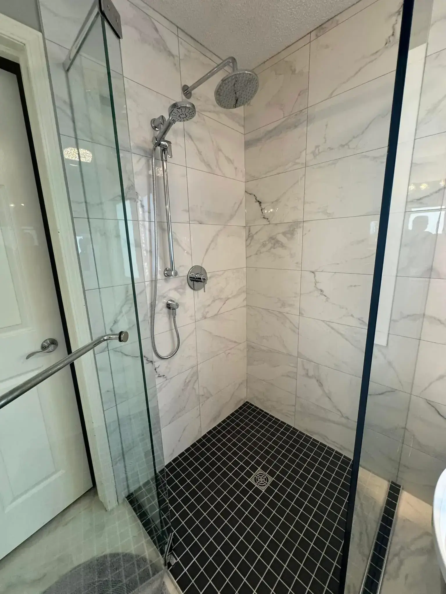 Walk-in shower with marble-look tile walls, black diamond mosaic floor, dual showerheads, and glass enclosure in Calgary