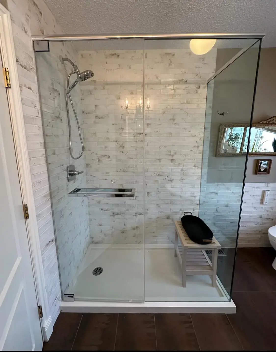 Frameless glass walk-in shower with textured brick-look tile, wooden bench, and dark hardwood flooring in Calgary bathroom