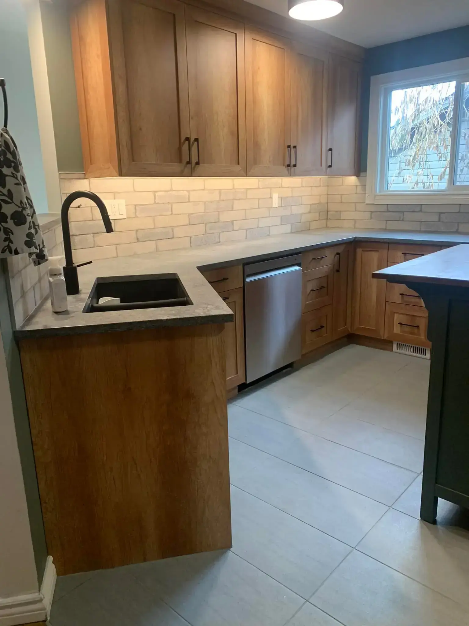 Kitchen with natural wood shaker cabinets, brick-look backsplash, black faucet, stainless dishwasher, and large tile flooring in Calgary